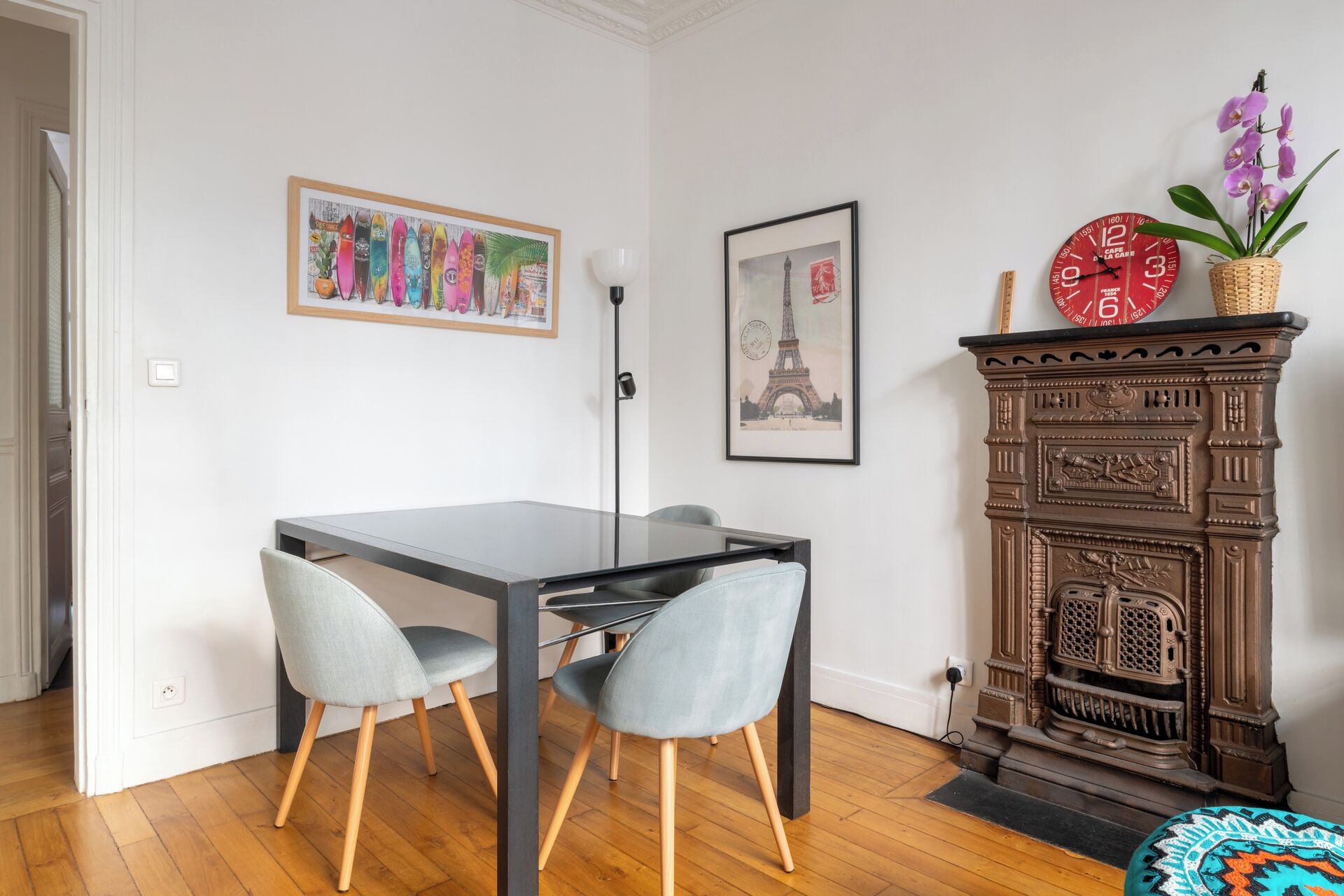 Un comedor con una mesa y sillas, una obra de arte enmarcada de tablas de surf, una fotografía enmarcada de la Torre Eiffel y una chimenea de estilo vintage con un reloj.