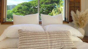 Cama com almofadas brancas e um manta estampada, janela com persianas de madeira e vista para vegetação exuberante.