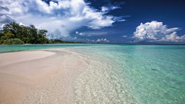 Uma praia com águas turquesa cristalinas, areia dourada e uma ilha coberta por vegetação ao fundo.