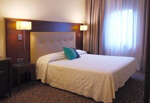 A hotel room with a large bed, two bedside lamps, a headboard, a nightstand, and a window with curtains. - Hotel Castello (Modena)