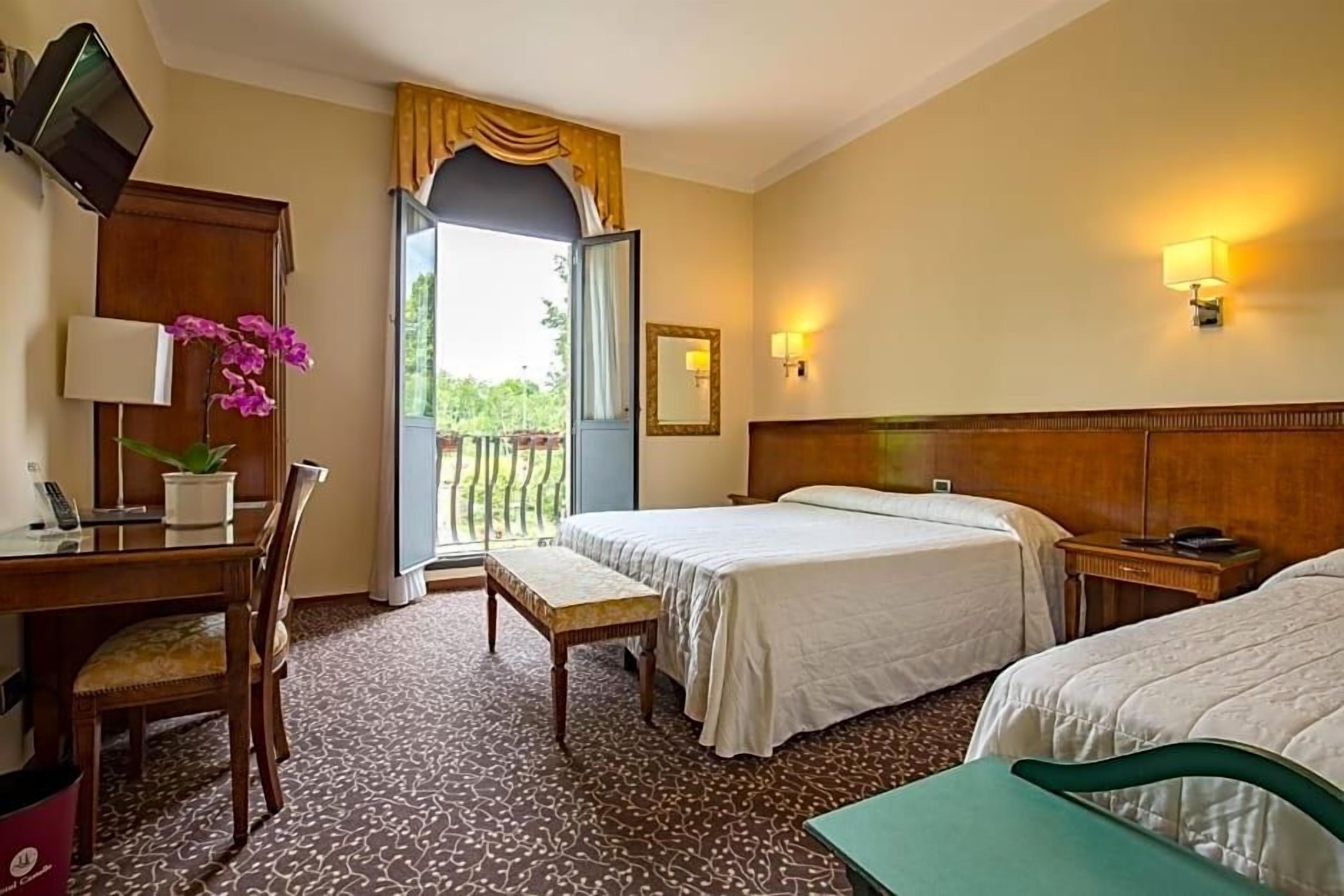 A hotel room with two beds, a desk with a lamp, a chair, a small table, a potted plant, and a balcony view.