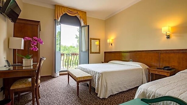 A hotel room with two beds, a desk with a lamp, a chair, a small table, a potted plant, and a balcony view. - Hotel Castello (Modena)
