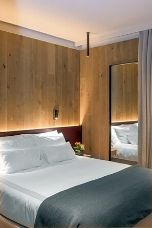 A modern hotel room with a large bed, wooden panel walls, a nightstand, and a mirror. - Olangerhof Hotel Spa (Valdaora)