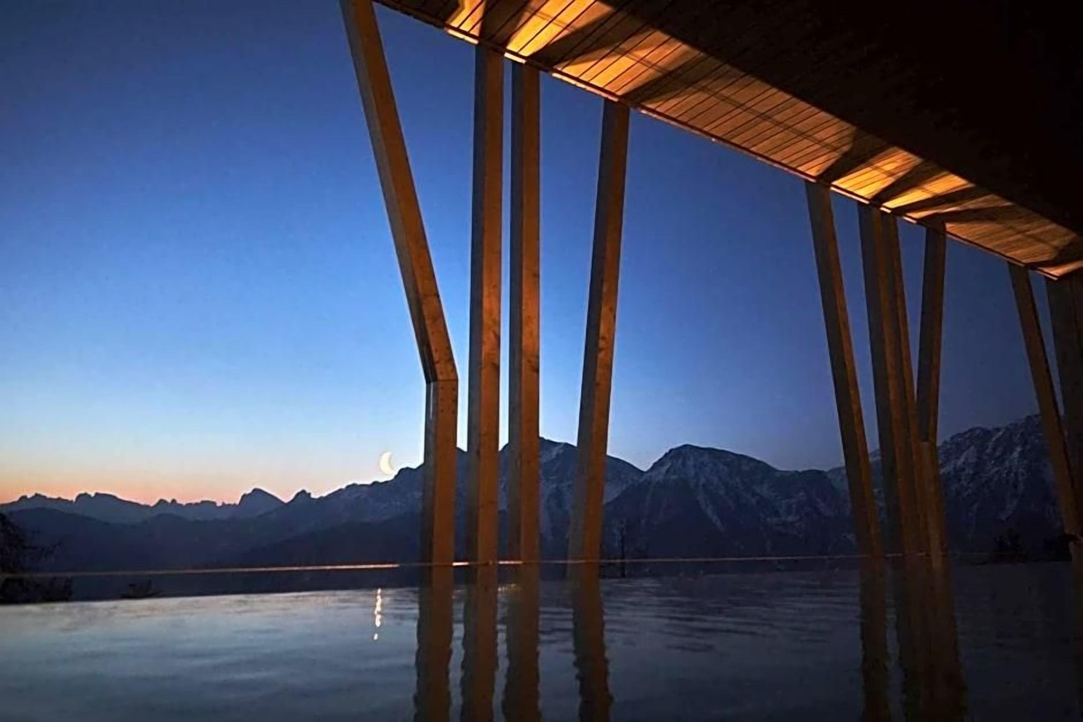 A wooden structure with a roof over a body of water, reflecting the surrounding mountains.