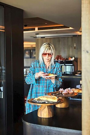 A person in a plaid shirt holding a plate with food, standing in a modern kitchen with wooden tables and a black countertop. - Olangerhof Hotel Spa (Valdaora)