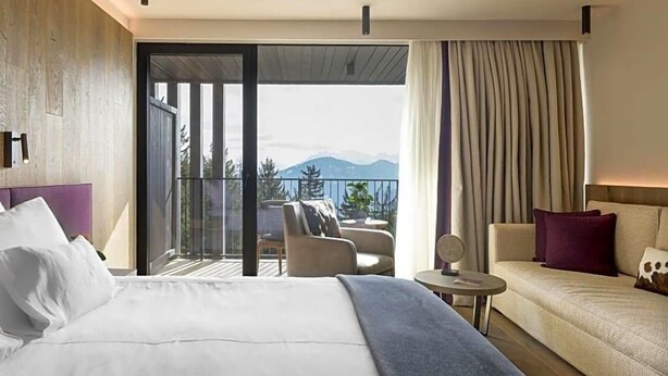 A hotel room with a bed, a sofa, a small table, and a balcony with a view of mountains. - Olangerhof Hotel Spa (Valdaora)