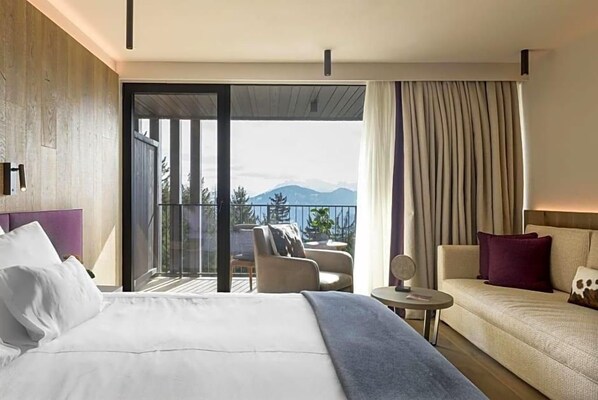 A hotel room with a bed, a sofa, a small table, and a balcony with a view of mountains. - Olangerhof Hotel Spa (Valdaora)