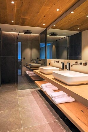 A modern bathroom with wooden ceiling, multiple sinks, and a long bench with towels. - Olangerhof Hotel Spa (Valdaora)