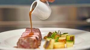 A white plate with a steak, vegetables, and a gravy boat pouring sauce. - Olangerhof Hotel Spa (Valdaora)