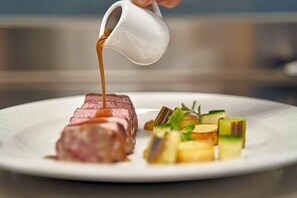 A white plate with a steak, vegetables, and a gravy boat pouring sauce. - Olangerhof Hotel Spa (Valdaora)