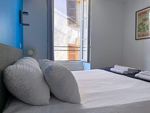 A bedroom with a bed, pillows, a fan, a window with blinds, and a framed picture on the wall. - Hostel 20 Bayonne (Bayonne)