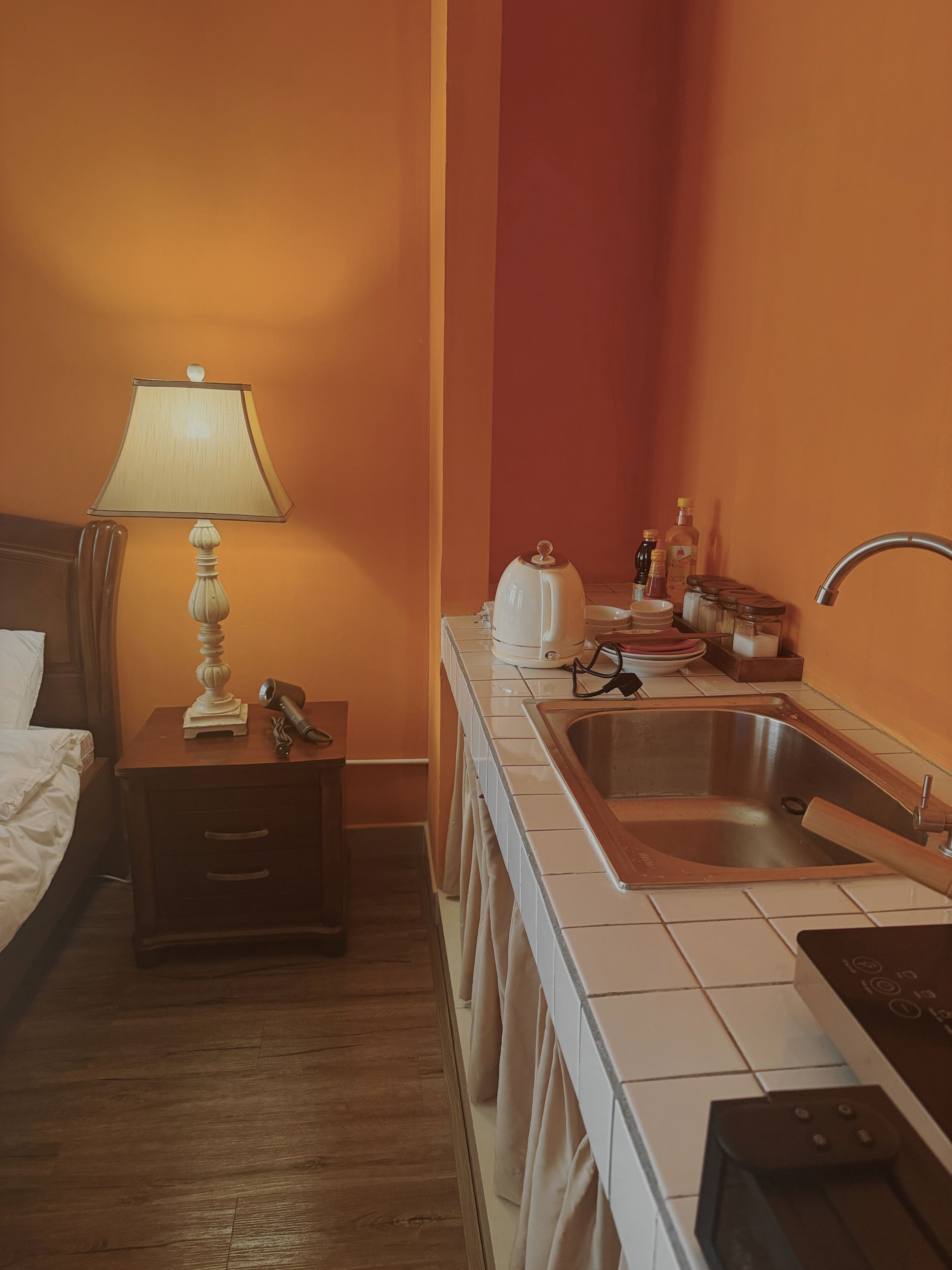 Habitación de hotel con cama, mesita de noche con lámpara, y una cocina pequeña con lavabo y armarios.