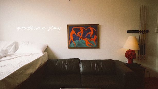 Quarto com cama, sofá de couro preto, pintura, luminária e um suporte na parede com gravetos.