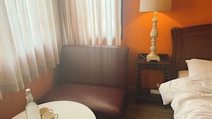 Quarto de hotel com cama, uma cadeira, uma mesinha com garrafa e copos, abajur e cortinas.