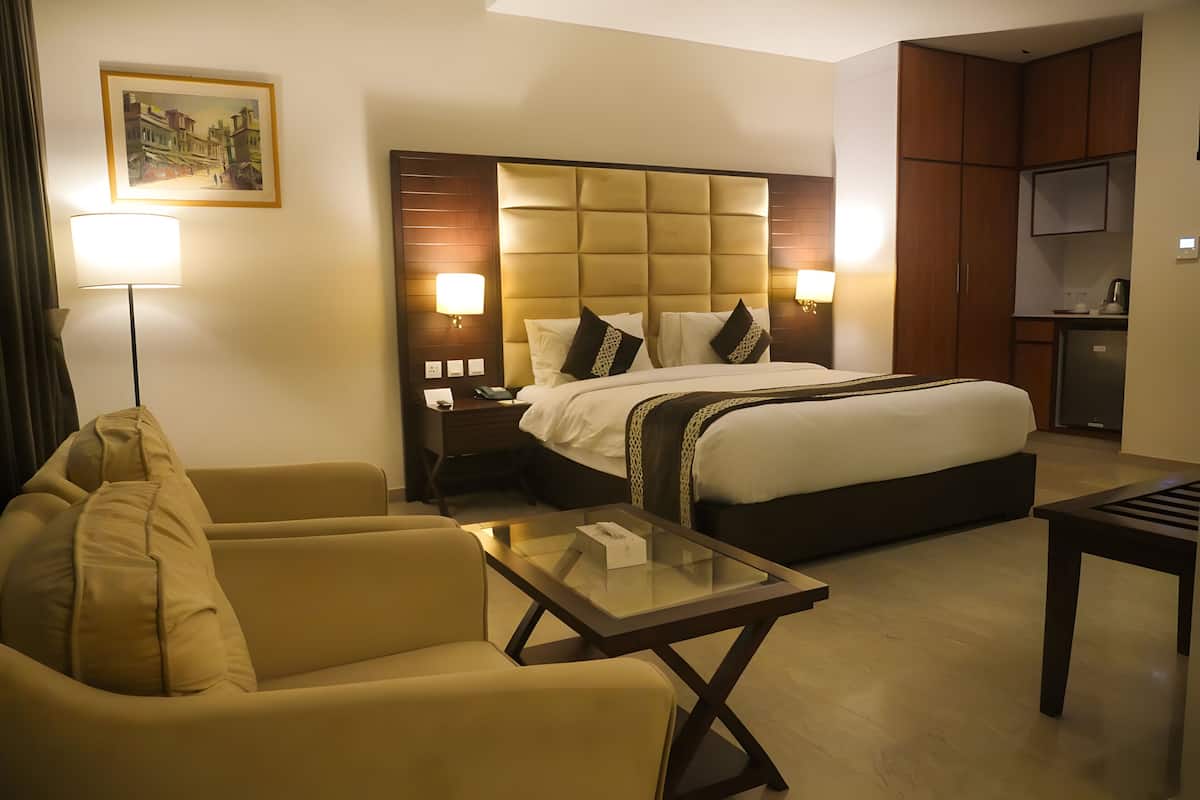 A hotel room with a bed, a sofa, a glass coffee table, a desk, a lamp, a TV, and a painting on the wall.