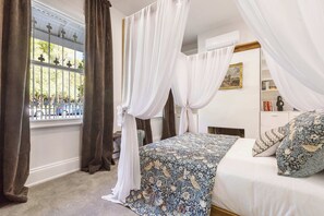 Ein Schlafzimmer mit Himmelsbett, einem Fenster mit Vorhängen, einem Nachttisch und einem Bücherregal.