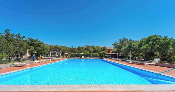 Ein rechteckiger Pool mit klarem, blauem Wasser, umgeben von einer gefliesten Liegefläche mit Liegestühlen und Sonnenschirmen, mit Bäumen im Hintergrund.
