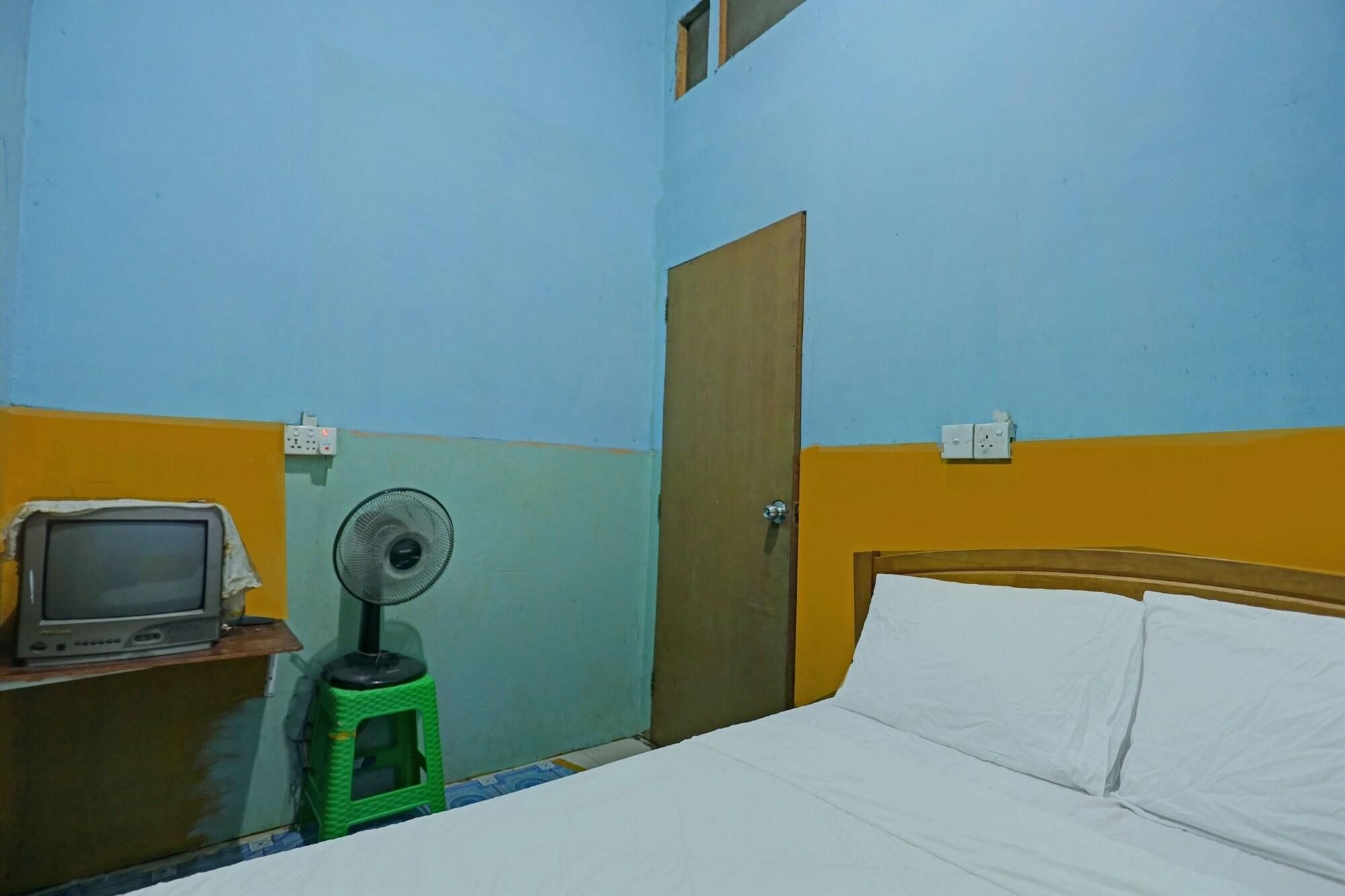 A room with a bed, a wooden headboard, a green stool with a fan, a television on a wooden table, and a door.
