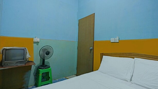 Ein Zimmer mit einem Bett, einem Holzkopfstück, einem grünen Hocker mit einem Ventilator, einem Fernseher auf einem Holztisch und einer Tür.