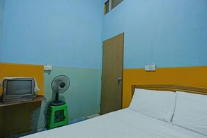 A room with a bed, a wooden headboard, a green stool with a fan, a television on a wooden table, and a door. - Spot On 92985 Mitra Homestay (Batam)
