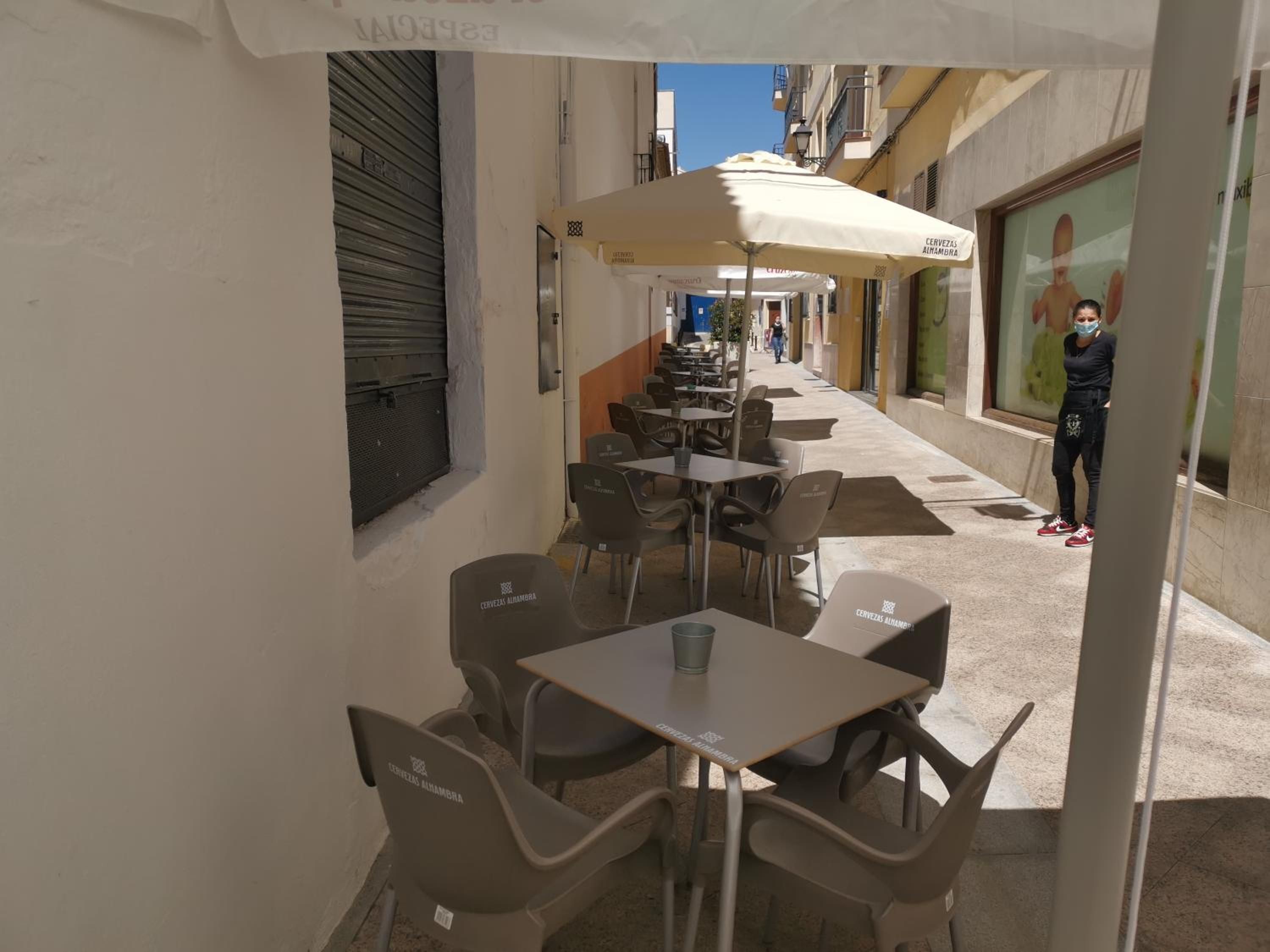 Un café al aire libre con mesas y sillas, un cerrojo en la izquierda y una persona cerca de la entrada.