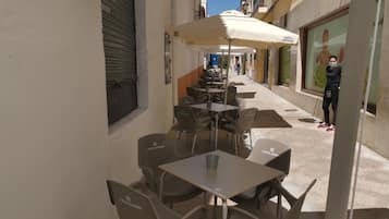 Un café al aire libre con mesas y sillas, un cerrojo en la izquierda y una persona cerca de la entrada.