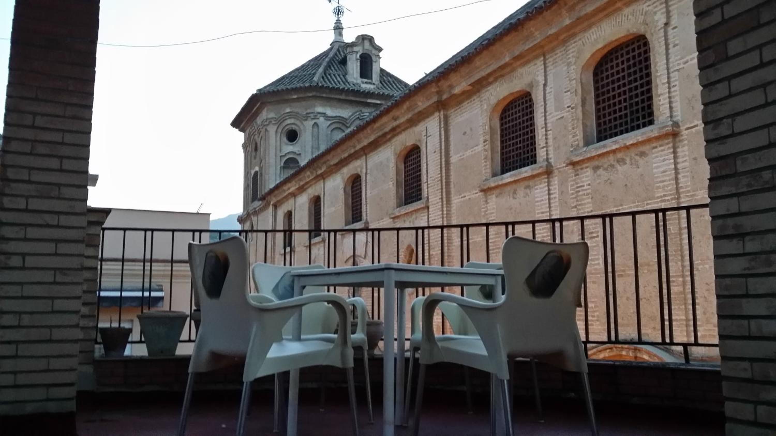 Un espacio al aire libre con mesas y sillas, un edificio de ladrillo con ventanas de arco y una torre de campana al fondo.