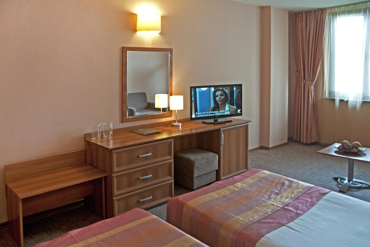A hotel room with a desk, a television, a bed, a chair, and a window with curtains.