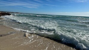 Uma praia com ondas, céu claro e uma linha de costa distante.