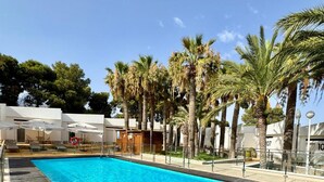 A swimming pool with a wooden deck, surrounded by palm trees and a white building. - El Eden (El Ejido)