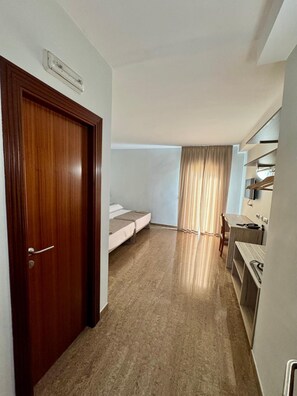 Une pièce de taille modeste, dotée d’une porte en bois, d’un lit, d’une petite kitchenette et d’une fenêtre avec des rideaux.