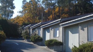 A row of single-story buildings with numbered garage doors, a paved road with directional arrows, and a backdrop of trees with autumn foliage. - Motel Cancun Barbadas Ourense (Barbadas)