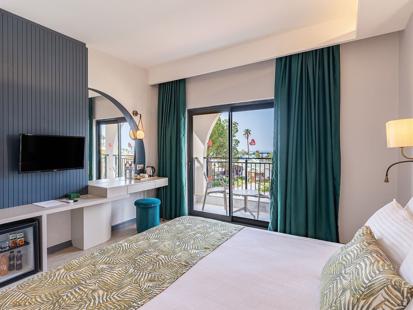 A hotel room with a bed, a TV, a desk with a mirror, a window with curtains, and a balcony with outdoor furniture.