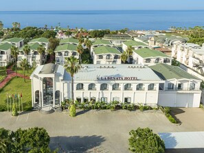 Een hotel met een centraal gebouw met de tekst 'La Benata Hotel', omringd door kleinere gebouwen met groene daken, palmbomen en uitzicht op zee.