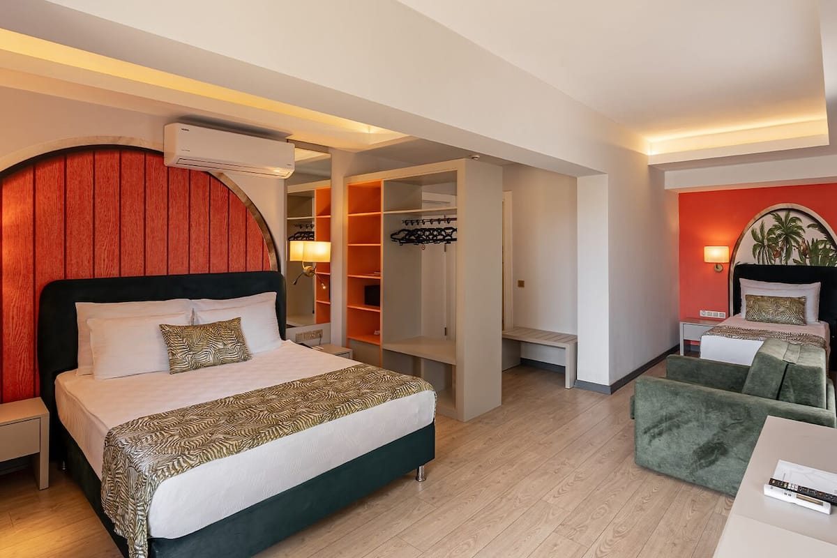 Hotellihuone, jossa on sänky, työpöytä, vihreä nojatuoli, vaatekaappi ja punaisella värillä korostettu seinä.