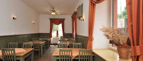 A restaurant with wooden tables and chairs, a ceiling fan, and a window with curtains.