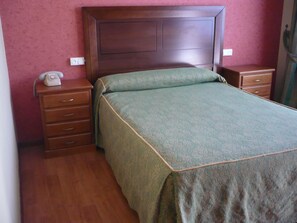 Un dormitorio con cabecera de madera, colcha verde, mesita de noche con teléfono y cómoda de madera.