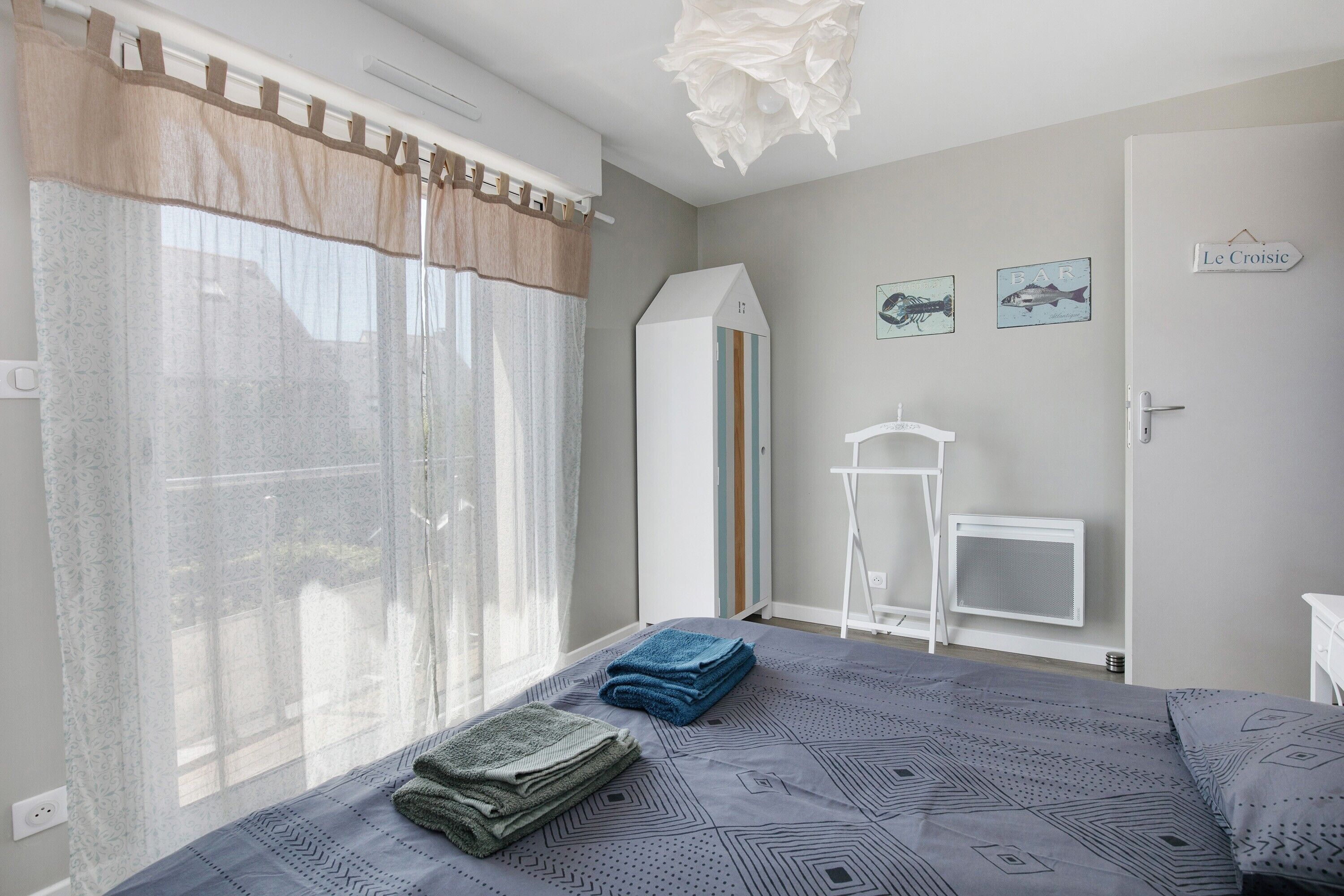 Une chambre à coucher avec un lit, une fenêtre avec des rideaux, un radiateur, une armoire blanche et une petite table blanche.