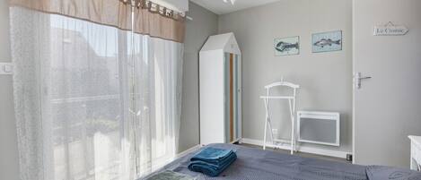A bedroom with a bed, a window with curtains, a radiator, a white wardrobe, and a small white table.