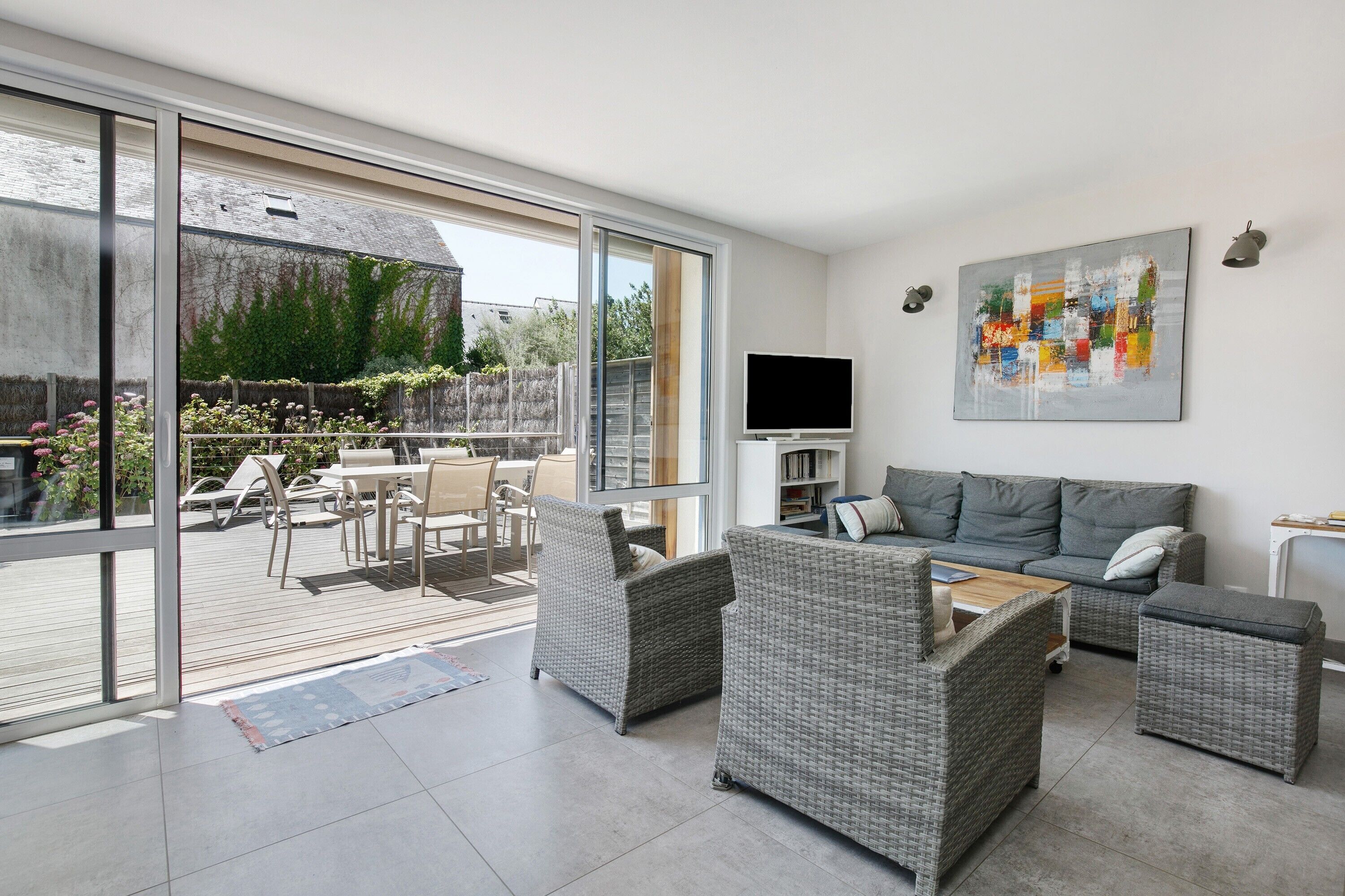 A modern living room with a large sliding glass door leading to an outdoor patio, a grey sofa set, a flat-screen TV, and a colorful abstract painting on the wall.