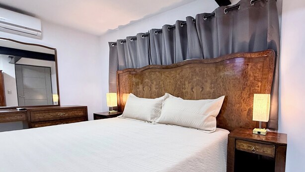 Ein Hotelzimmer mit einem großen Bett, einem Holzkopfteil, zwei Nachttischen mit Lampen, einem Spiegel und einer Klimaanlage.