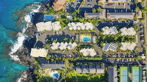 Eine Vogelperspektive zeigt einen Küstenort mit mehreren bungalowsartigen Unterkünften, einem zentral gelegenen Pool und einem Tennisplatz.