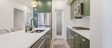 A kitchen with green cabinets, a white countertop, stainless steel appliances, and a patterned rug.