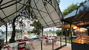 An outdoor dining area with tables and chairs, a covered seating area, and a view of a river. - Ayutthaya Rive Camp And Yacht Charter (Ayutthaya)