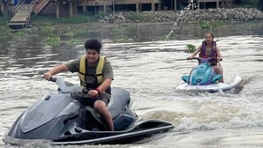 Two people on watercrafts in a body of water with a bridge and buildings in the background. - Ayutthaya Rive Camp And Yacht Charter (Ayutthaya)