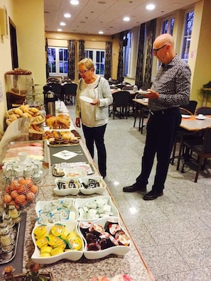 A buffet setup with various food items and a man and woman standing by the counter. - Hotel De Venne (Genk)