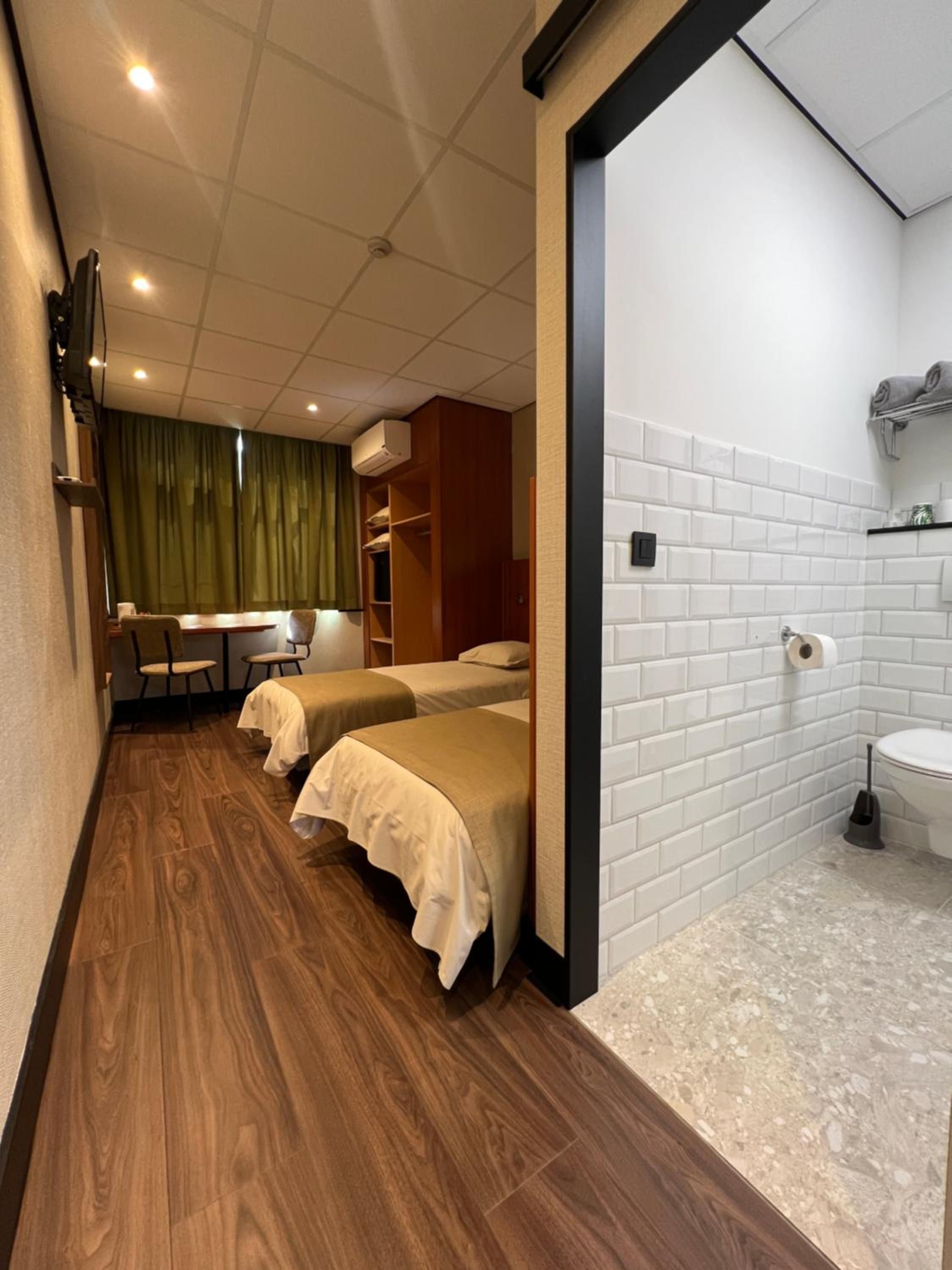 Een hotelkamer met een houten vloer, twee bedden, een klein bureau en een badkamer met witte tegels.