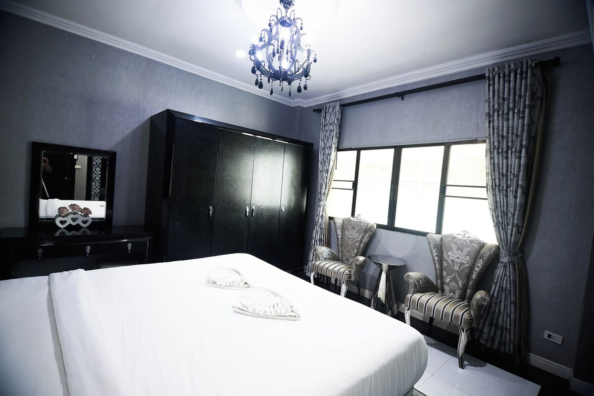 Een slaapkamer met een groot bed, een kledingkast, een stoel, een tafeltje en een raam met gordijnen.