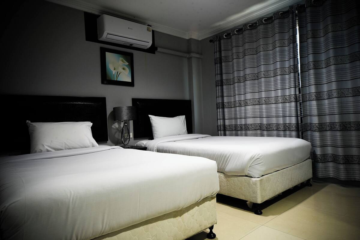 Hotellihuone, jossa on kaksi sänkyä, kattotuuletin, ilmastointilaite ja ikkuna, jossa on kuvioidut verhot.