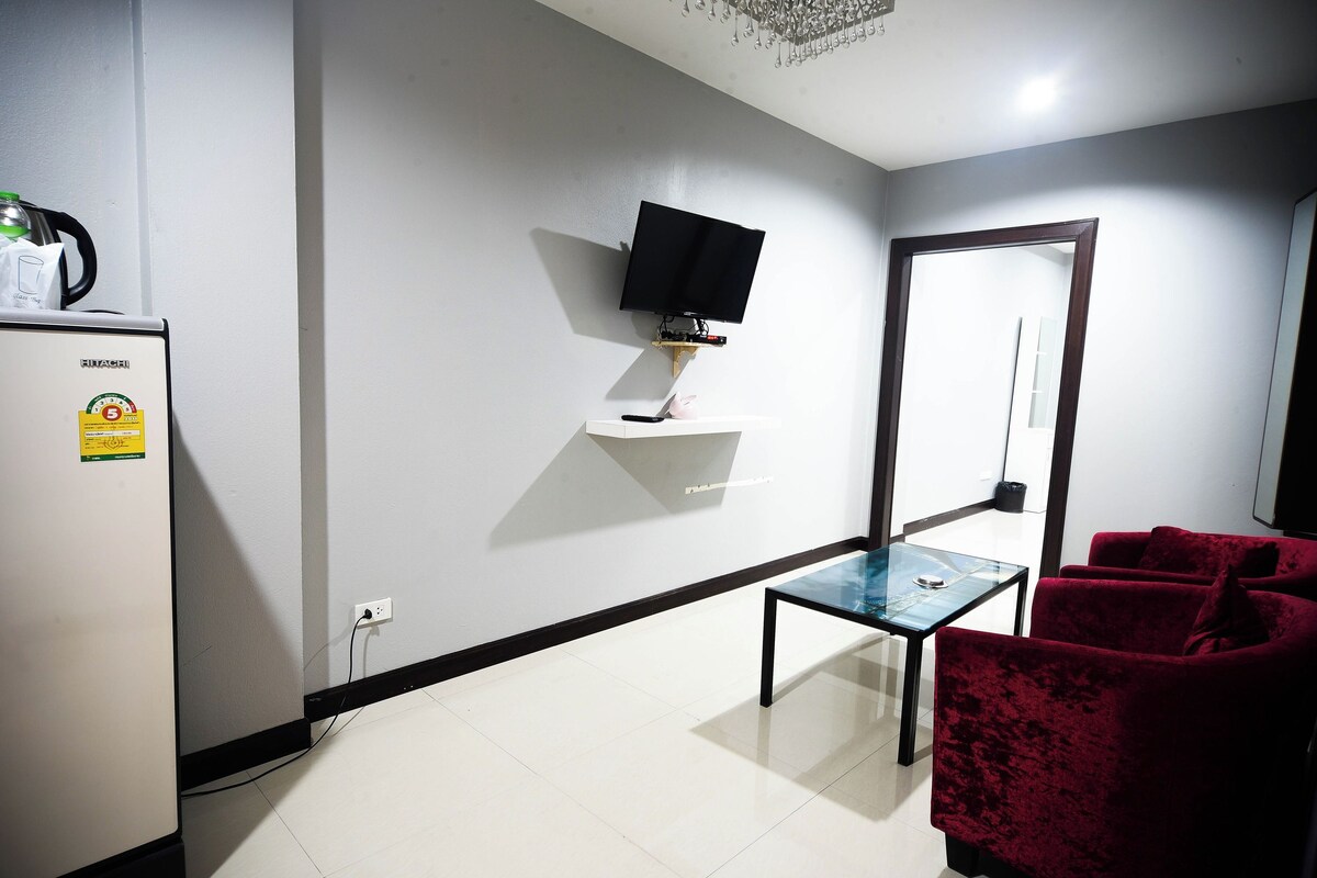A modern living room with a flat-screen TV mounted on the wall, a glass coffee table, a red sofa, and a refrigerator with a sticker.